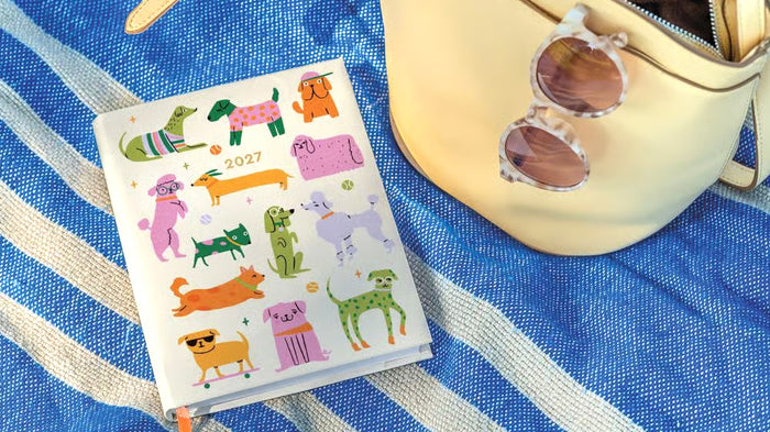 A planner with a white cover decorated with colorful cartoon dog illustrations and the year 2027, styled on a blue and white striped blanket next to a cream-colored bag with sunglasses.