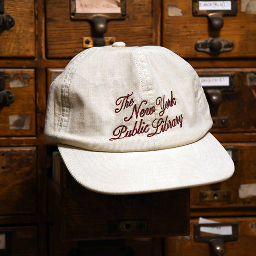 An off-white cotton baseball cap with 'The New York Public Library' embroidered in dark red script on the front, displayed on a wooden library card catalog drawer.